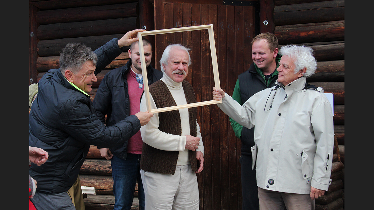 Artists posing with an empty picture frame outside a traditional log house.