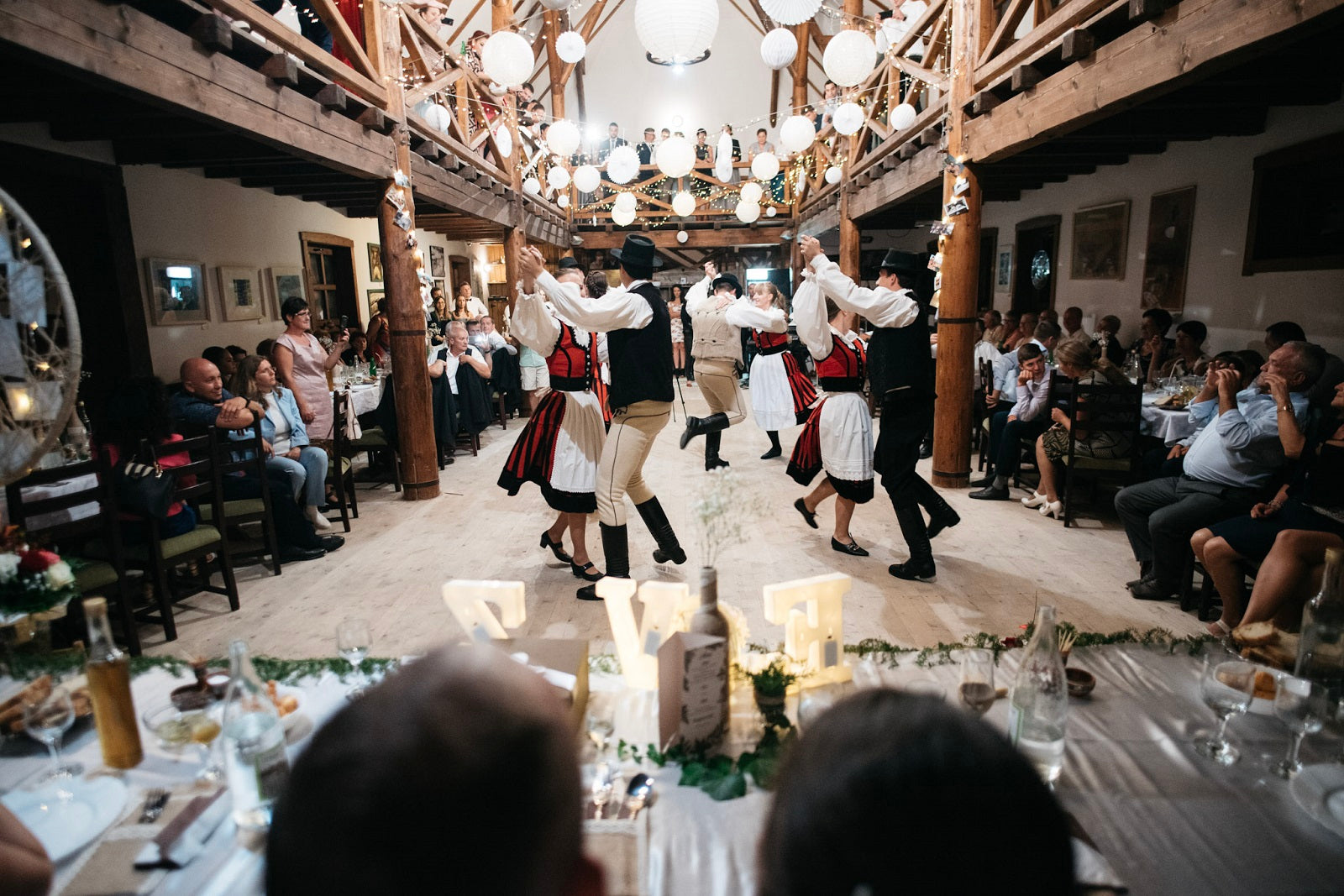Folk dancers holding hands in a circle inside the event barn, lit by hanging paper lanterns.