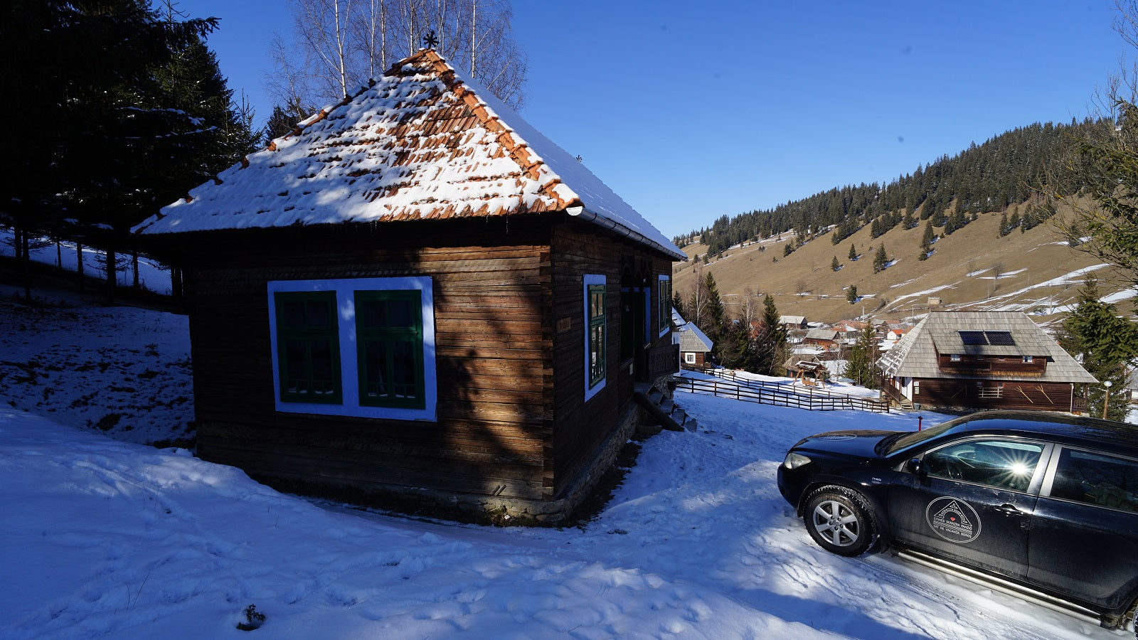 A snow-covered wooden house with a car parked outside, forested hills in the winter distance.