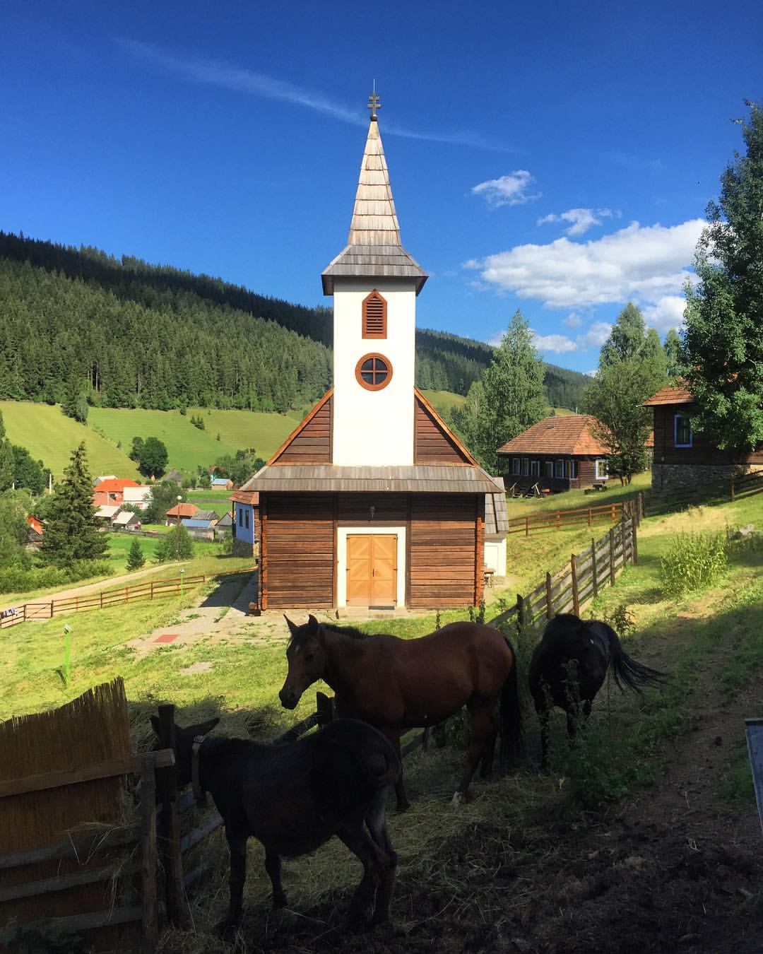 Horses graze in a fenced pasture in front of a wooden church with a tall shingled steeple.