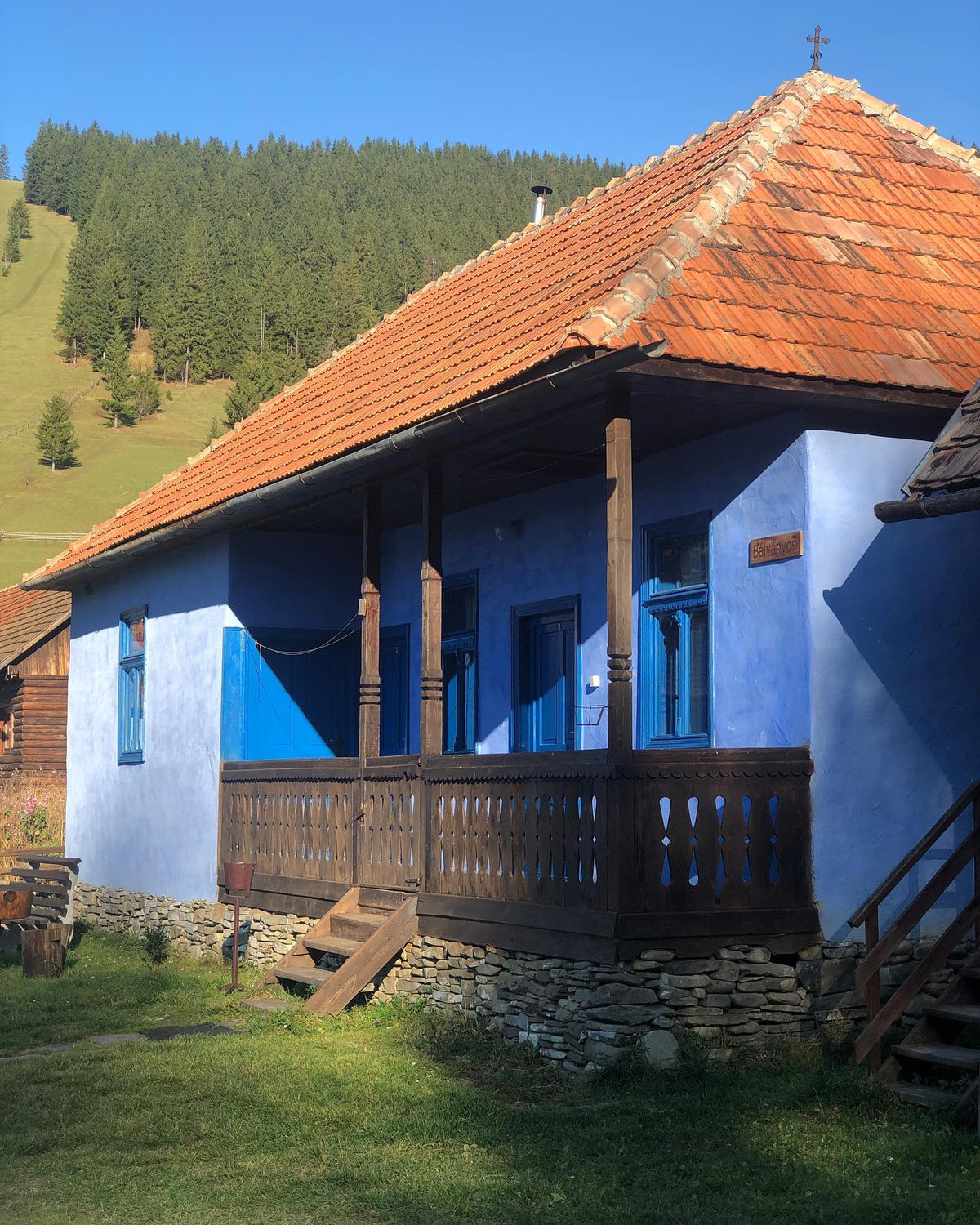 Bálványos house at ground level, with blue-framed windows and doors and a carved wood porch railing.