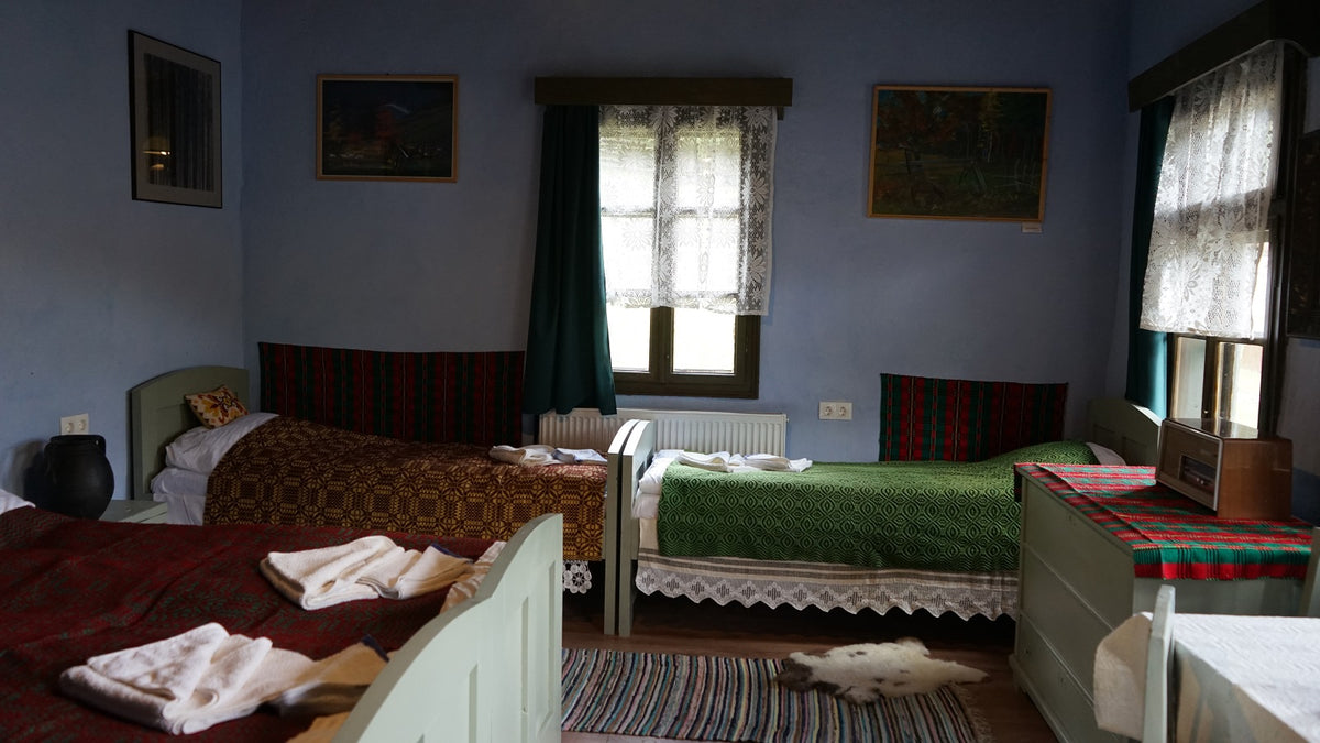 Multi-bed room with maroon, mustard, and green folk-pattern covers, lace-curtained windows, and framed landscape paintings.