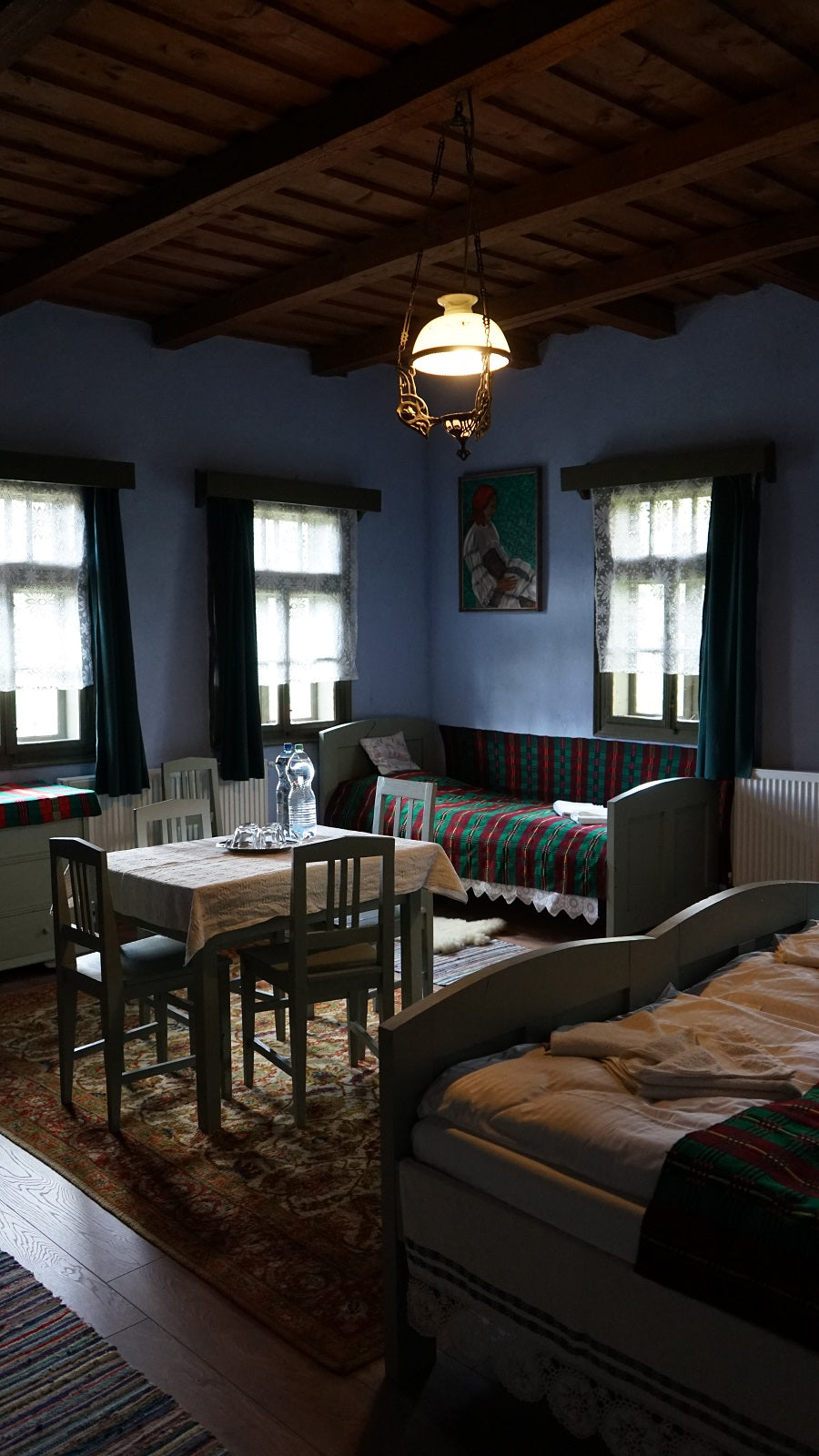 Blue bedroom with lace curtains, a folk-costume portrait painting, and beds dressed in red and green plaid.