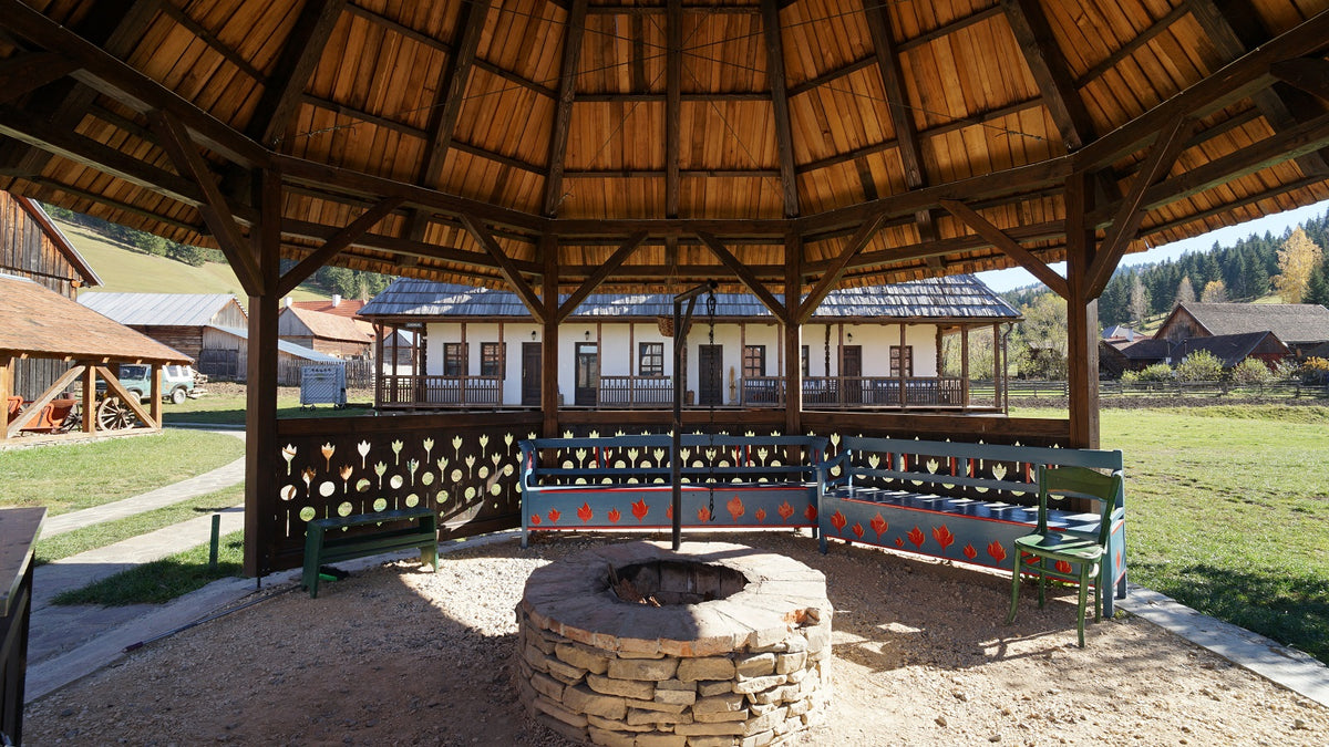 Octagonal wooden gazebo with a stone fire pit and colorful painted benches, the Csorja house behind it.