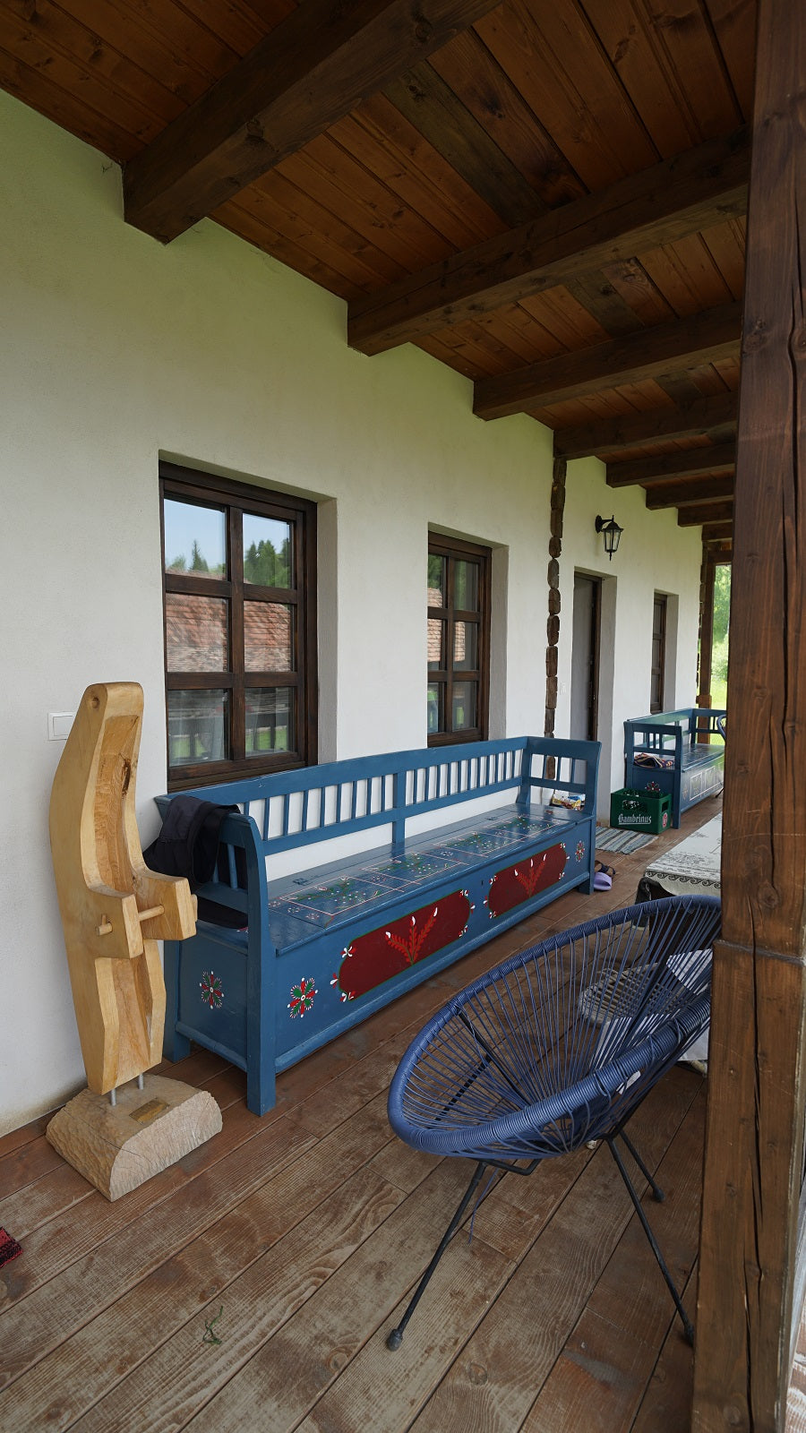 Covered porch with a blue folk-painted bench, a woven chair, and a carved abstract wood sculpture.