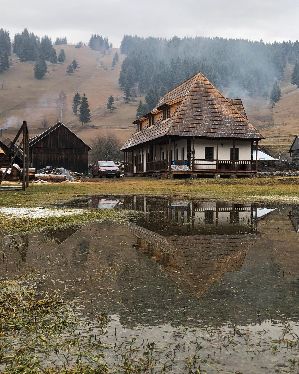 Csorja house reflected in a melting-snow puddle, with misty hills and a red car parked nearby.