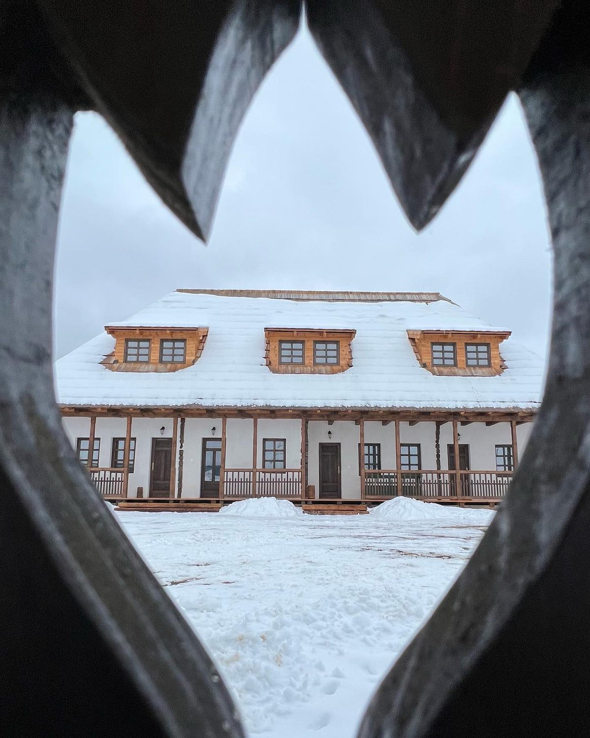 Snow-covered Csorja house framed through a heart-shaped cutout in the carved wooden fence.