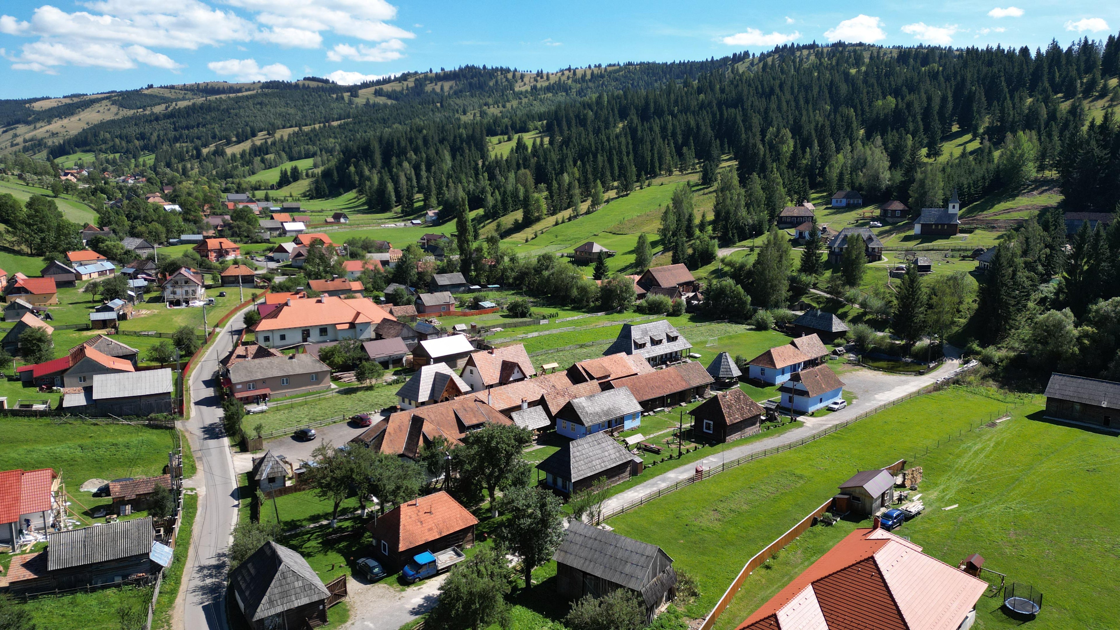Aerial view of a small mountain village with scattered houses, red-orange roofs, green fields, and dense evergreen forests under a blue sky with clouds.