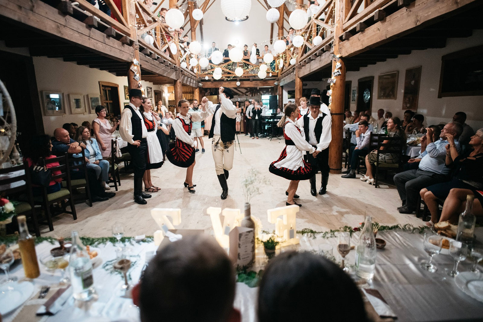 Folk dancers performing for wedding guests inside the event barn, paper lanterns hanging from the rafters.