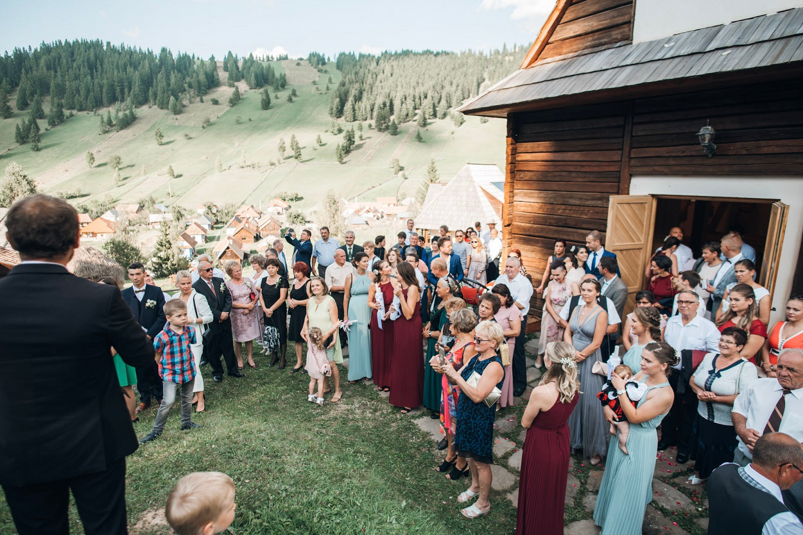 Wedding guests gathered outside the wooden chapel, hills and the village rooftops spreading below.