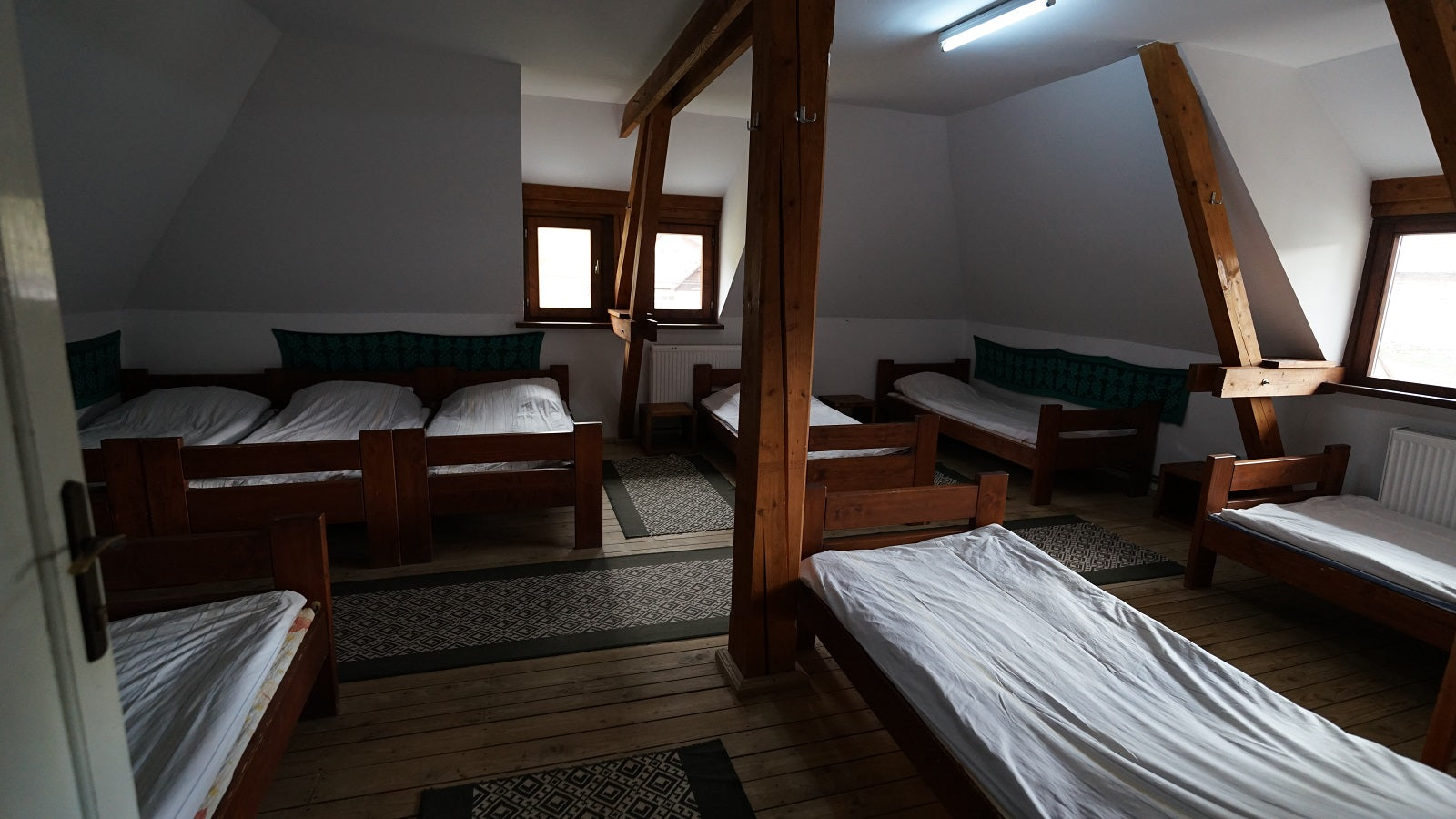 Larger attic dormitory with several wooden twin beds arranged around exposed roof-truss beams.