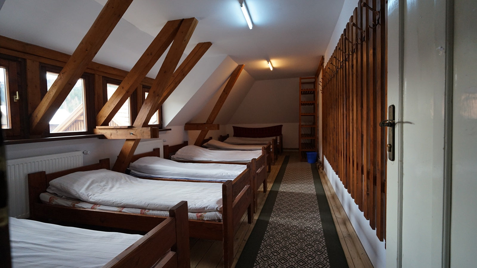 Long attic dorm room with a row of wooden twin beds beside a wall of wooden coat hooks.
