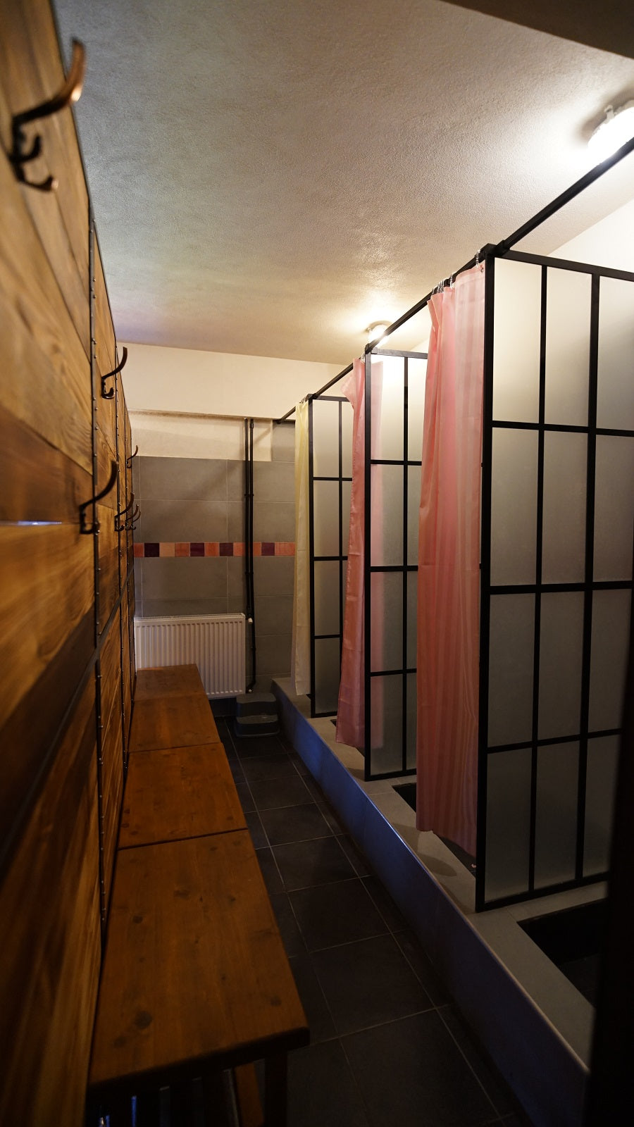 Changing area with wooden benches and coat hooks, beside a row of glass-partitioned shower stalls with striped curtains.