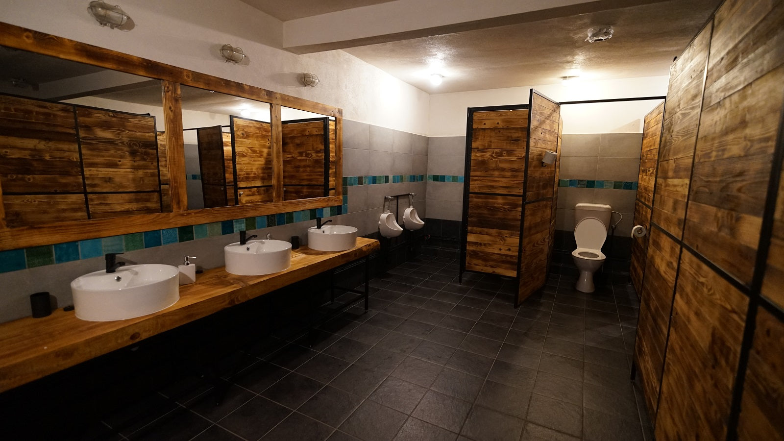 Men's washroom with vessel sinks, urinals, and wood-plank toilet stalls trimmed in teal tile.
