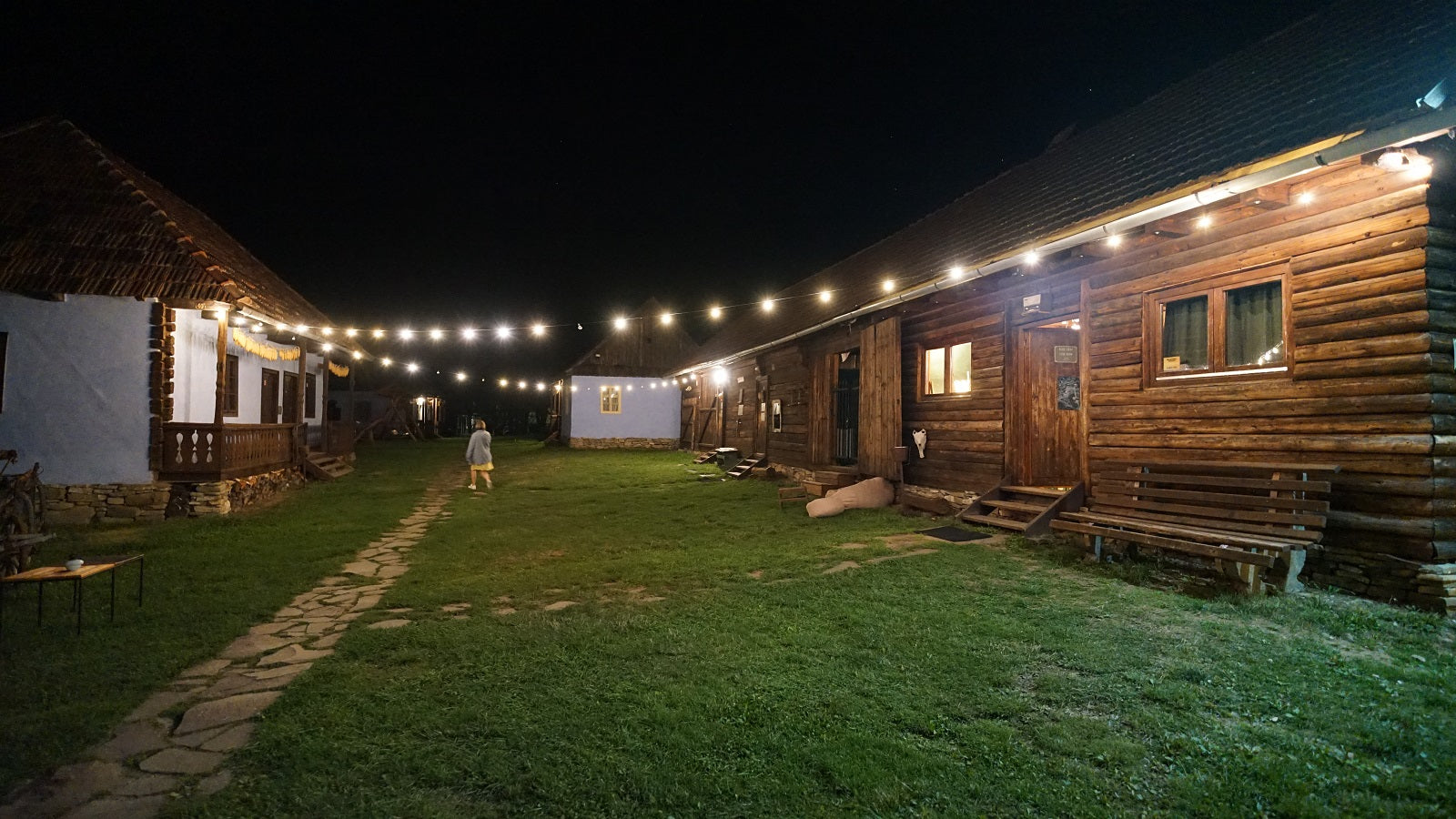 Museum grounds lit with string lights at night, log-cabin buildings glowing warm against the dark sky.