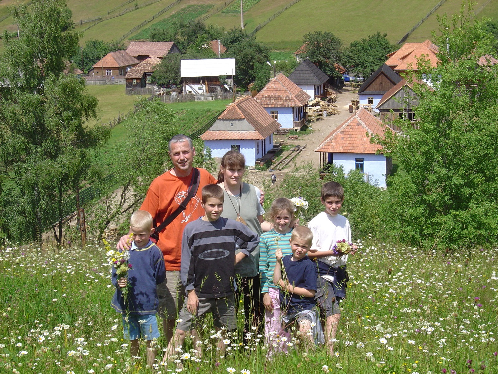 Family standing together in a wildflower meadow above the village, its half-finished houses visible below.