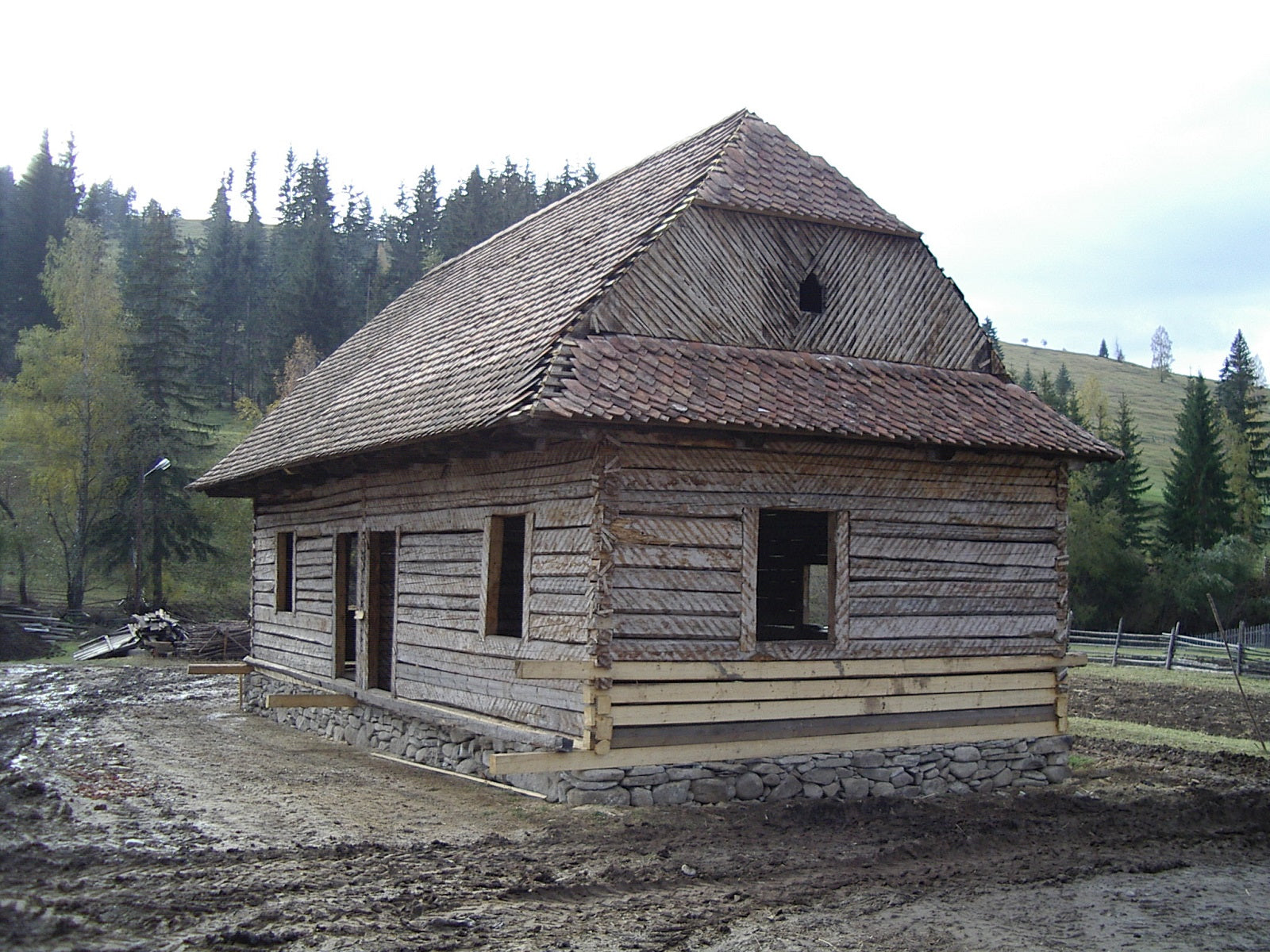 A log house mid-construction, its shingle roof finished but windows still open, framed by forested hills.