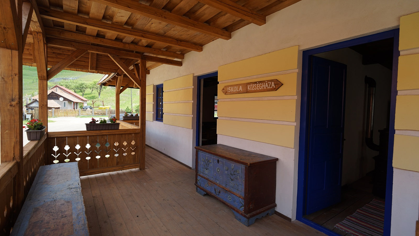 Porch signpost pointing to Iskola (School) and Községháza (Village Hall), beside a painted blue folk chest.