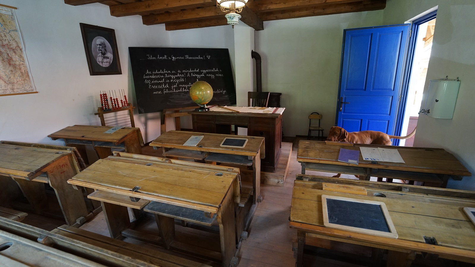 Classroom with wooden desks holding small slate boards, a cast-iron stove, and a dog sniffing near the open door.