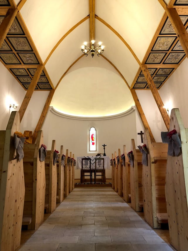 View toward the chapel altar with gray fabric bows on the pews, painted ceiling panels, and a hanging chandelier.