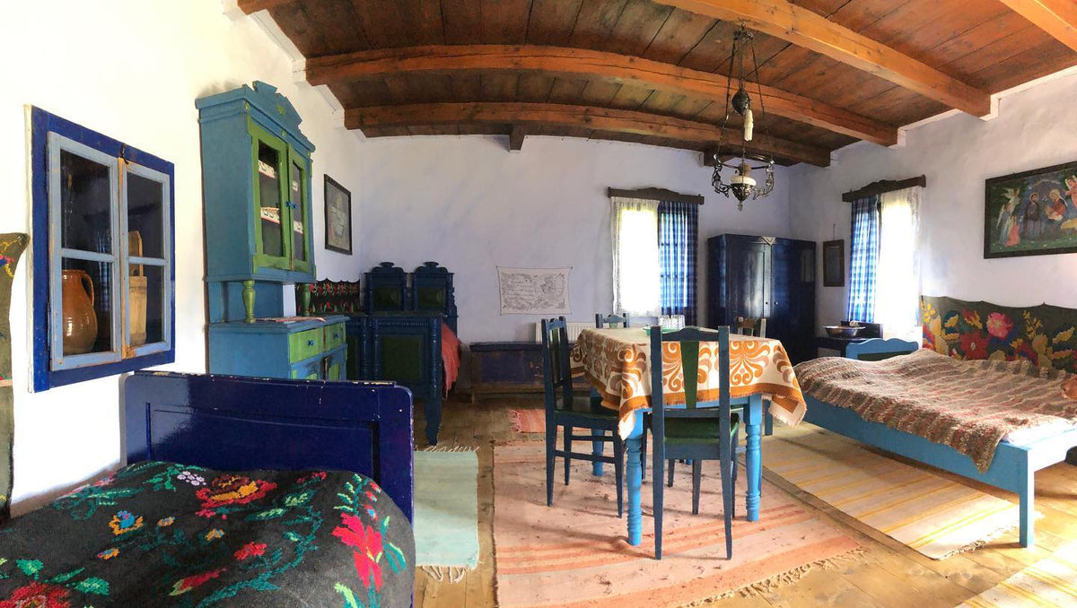 Room with dark blue and green painted antique furniture, floral folk-woven blankets, and an arched wood-beamed ceiling.
