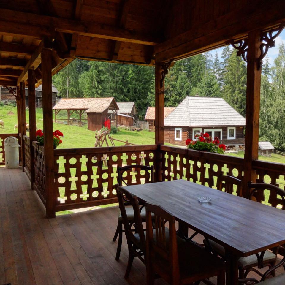 Covered wooden balcony with a dining table and carved cross-motif railing, overlooking the museum grounds and grazing sheep.