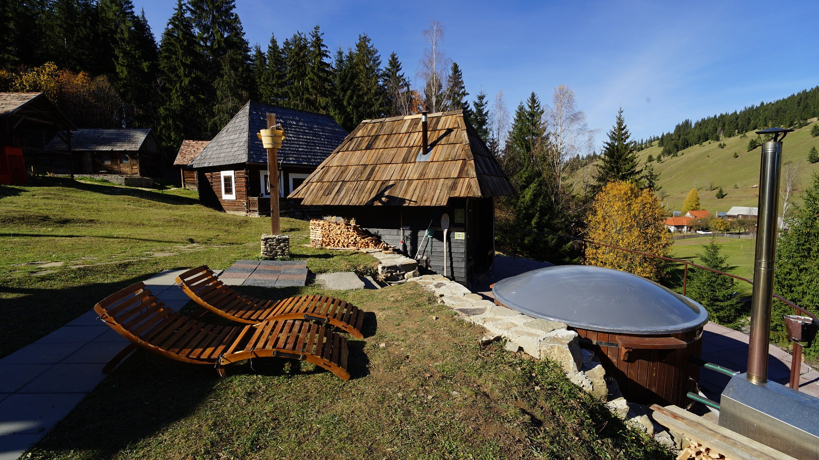 Wood-fired hot tub and a shingle-roofed sauna cabin on a terrace, with wooden lounge chairs and forested hills beyond.