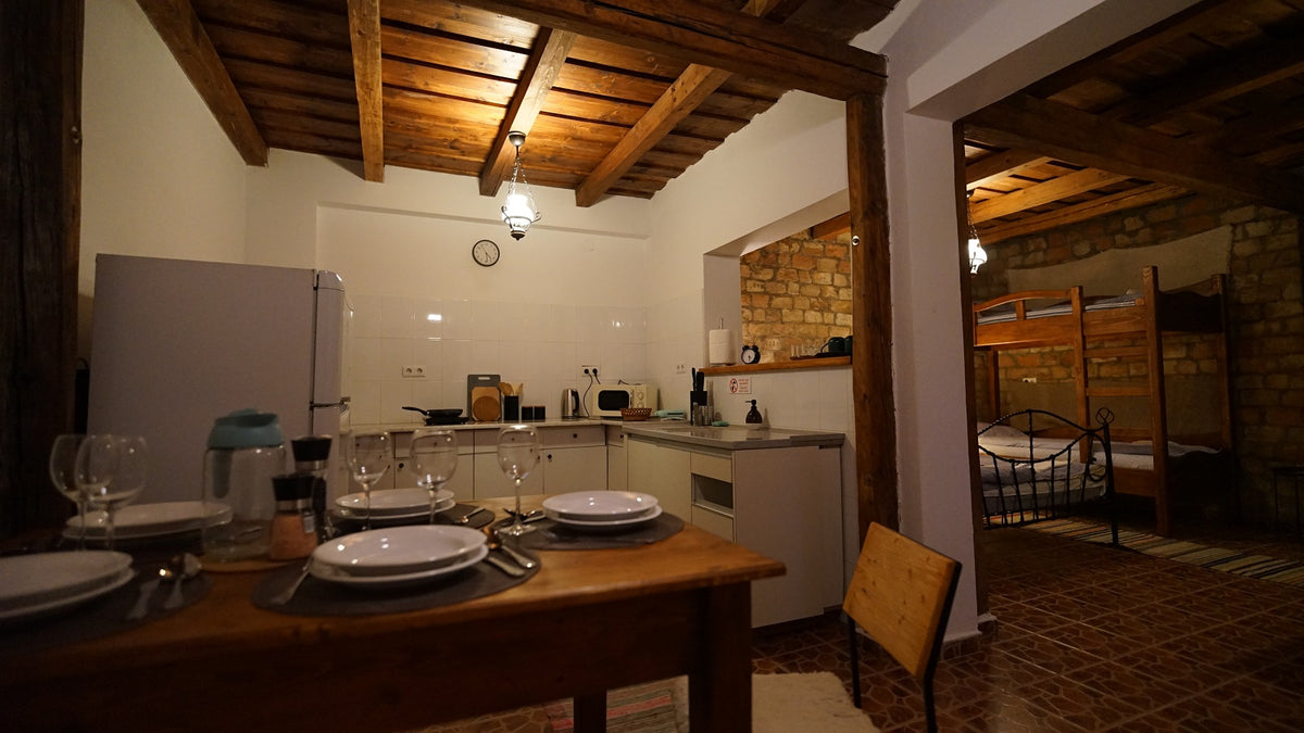 Kitchen with a set dining table and wooden beamed ceiling, opening through a brick archway into a bedroom with bunk beds.