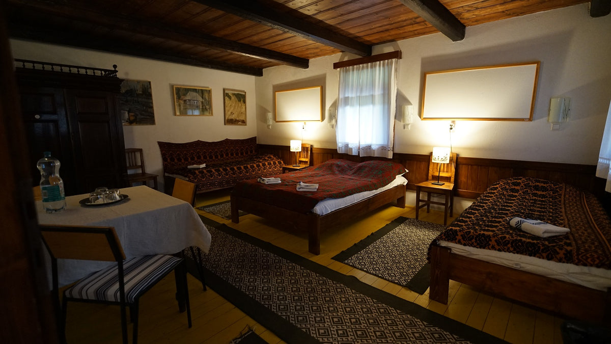 Room with three beds in dark folk-patterned covers under a wood-beamed ceiling, furnished with a small dining table.