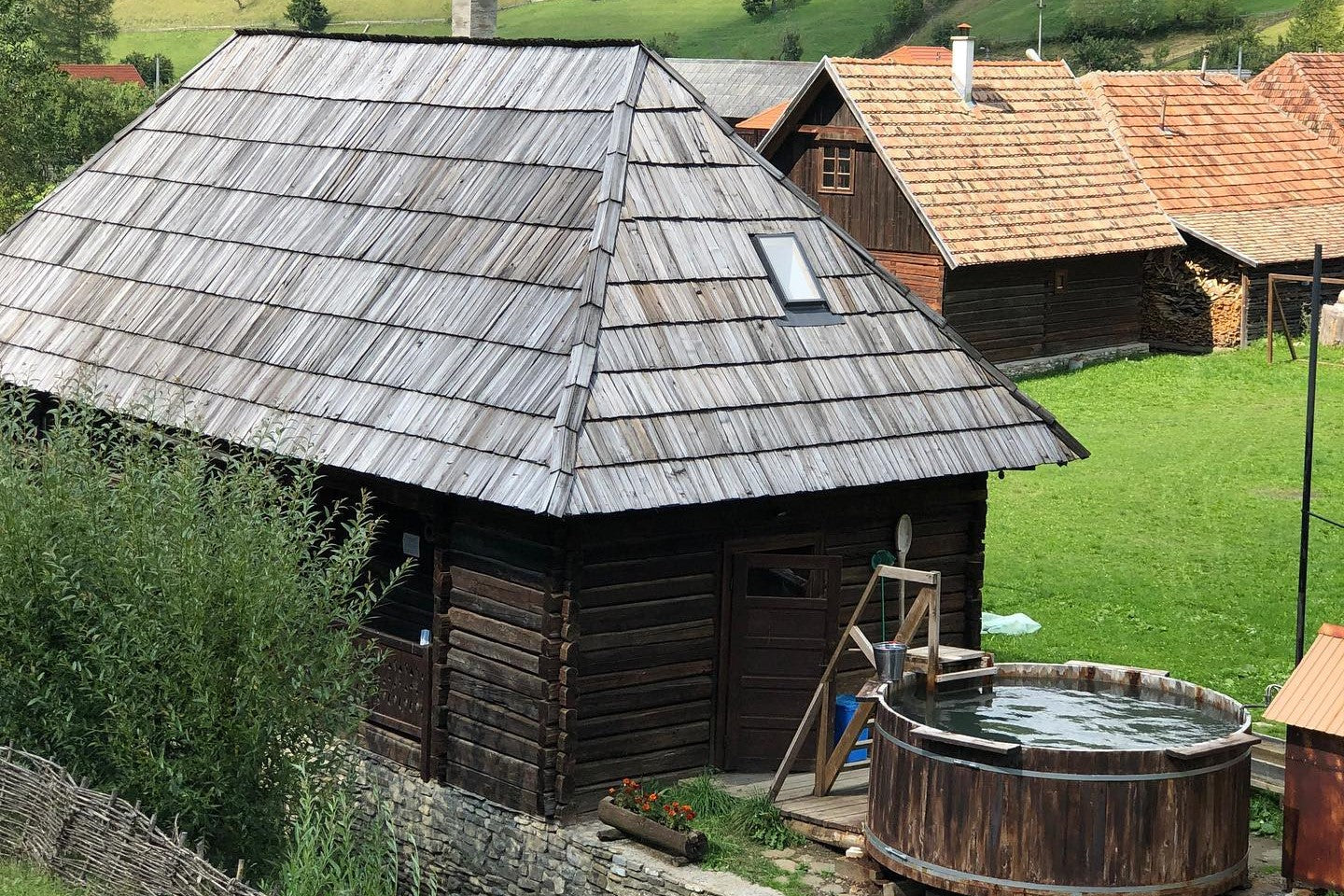Log-built mill and sauna house with a steep shingle roof and a water-filled wooden dézsa tub on the deck, hills behind.
