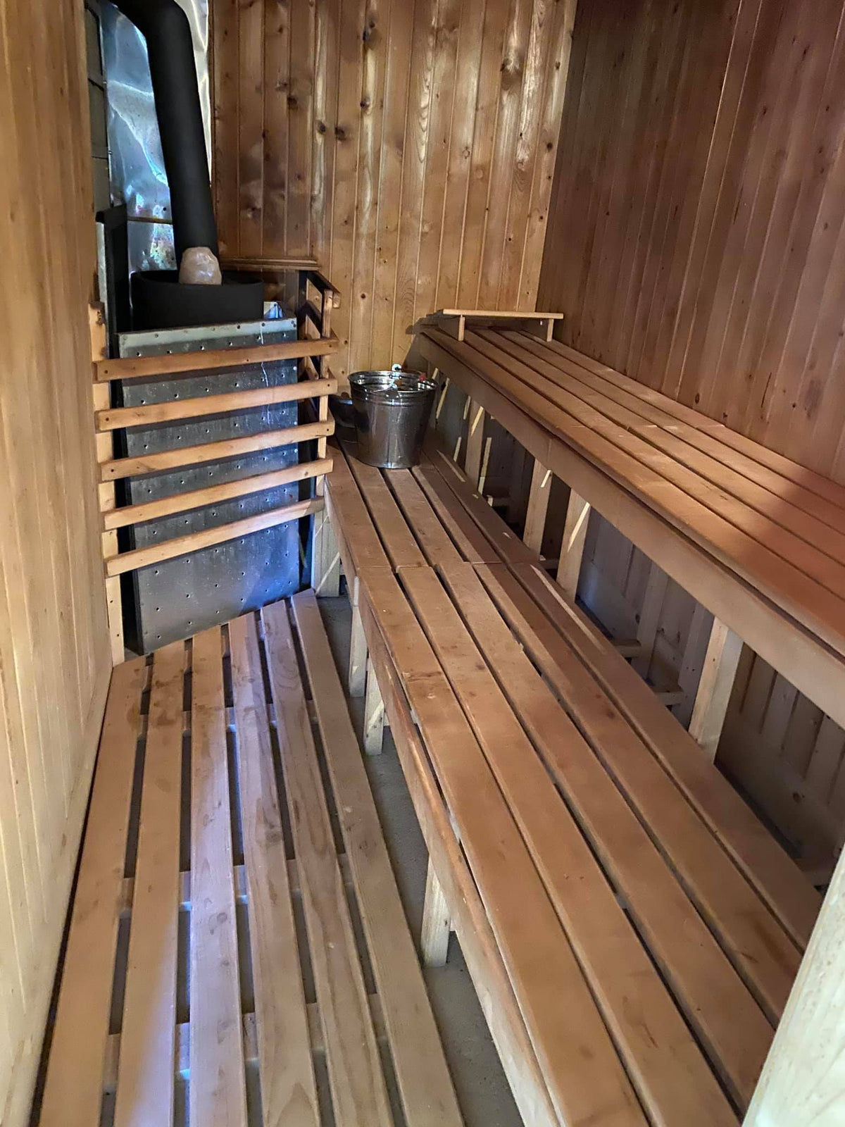 Interior of the sauna with tiered wooden benches, a black stove topped with stones, and a metal bucket.