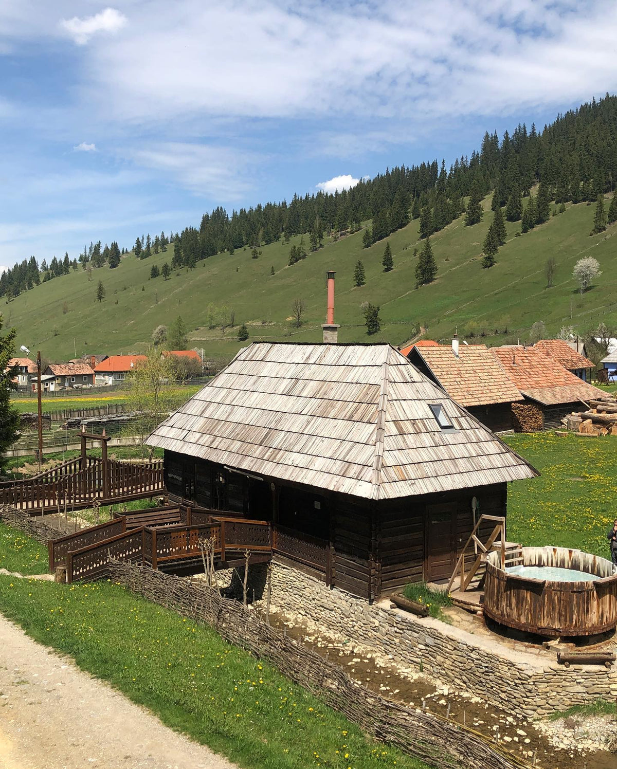 Sunny exterior view showing the woven wicker fence, the wooden tub on the deck, and hills rising behind.