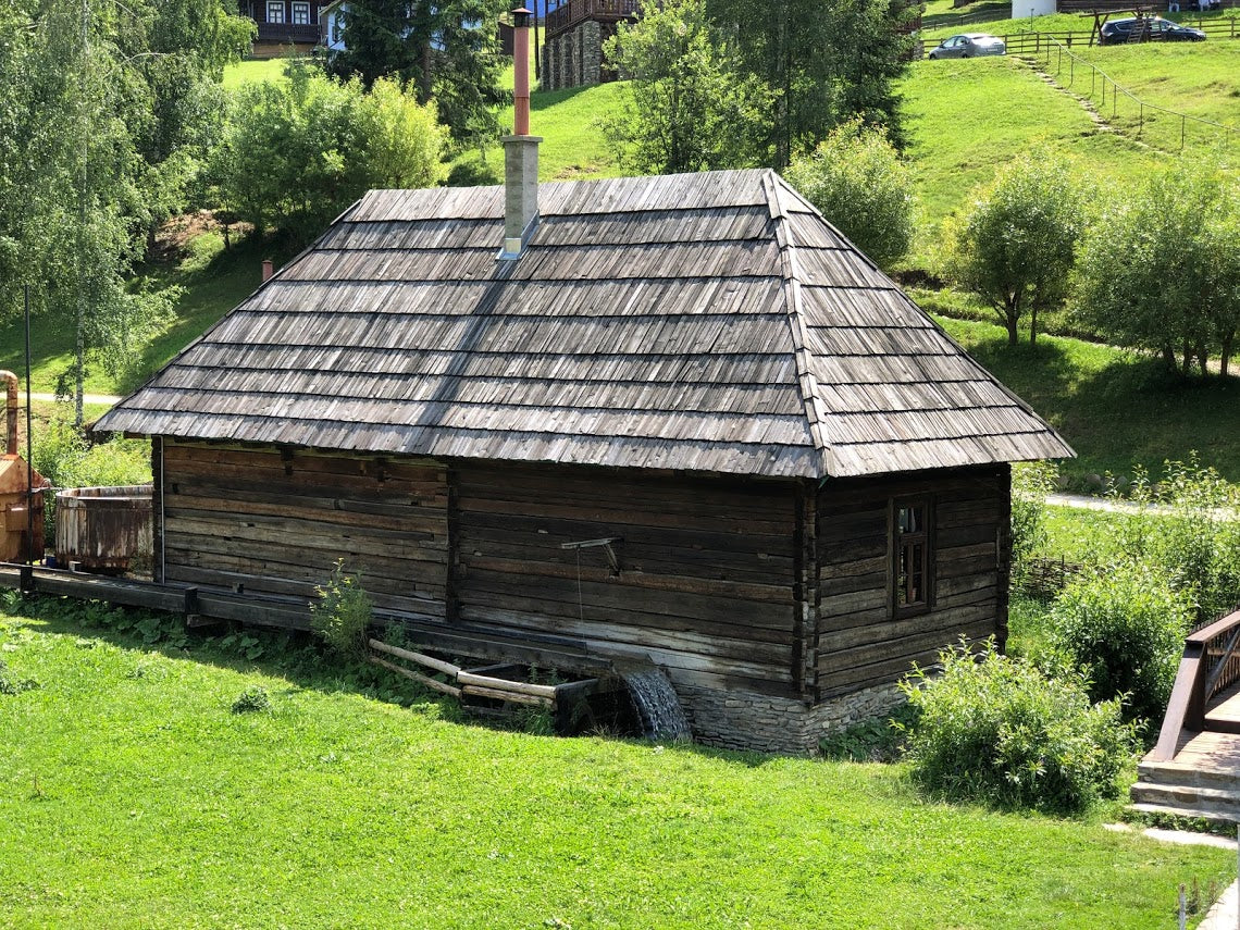 Side view of the building with an old wheel leaning against the log wall beside its water trough.