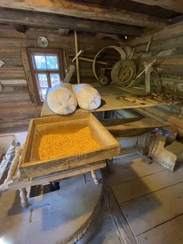 Interior of the mill with a wooden hopper of corn kernels above the millstone, sacks and gears nearby.