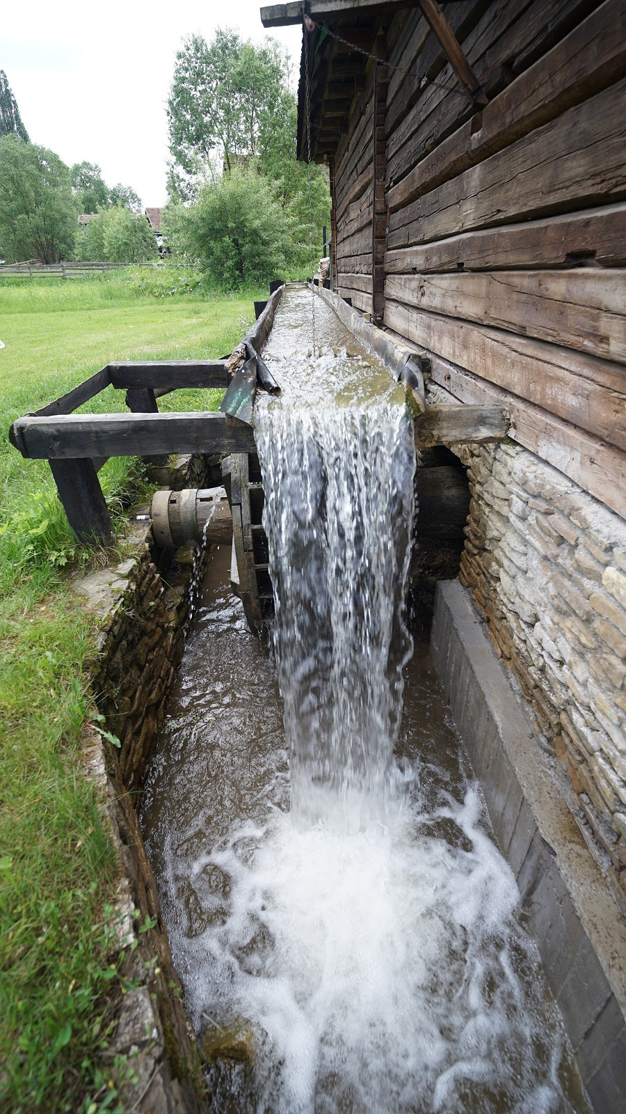 Water pours down a wooden trough into a stone-lined channel outside the mill, green field and trees behind.