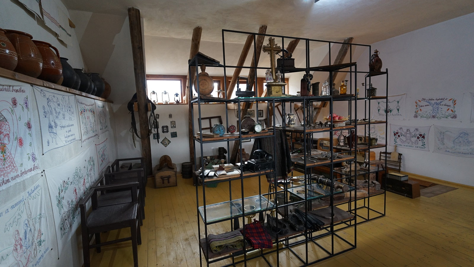 Attic exhibit room with clay jugs on a shelf, embroidered folk-poem cloths on the wall, and glass display shelving.