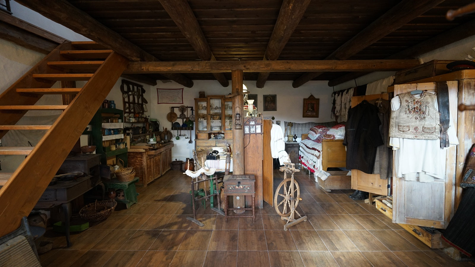 Museum room with a wooden staircase, an antique dresser, a spinning wheel, and folk costumes hanging from a rail.