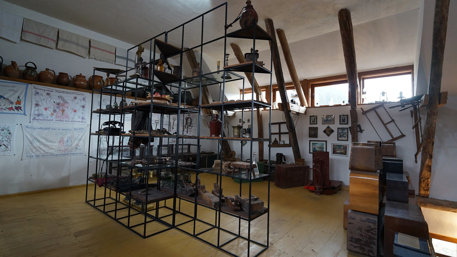 Attic display room with stacked leather trunks, a crucifix and clay jugs on glass shelving, beneath sloped beams.