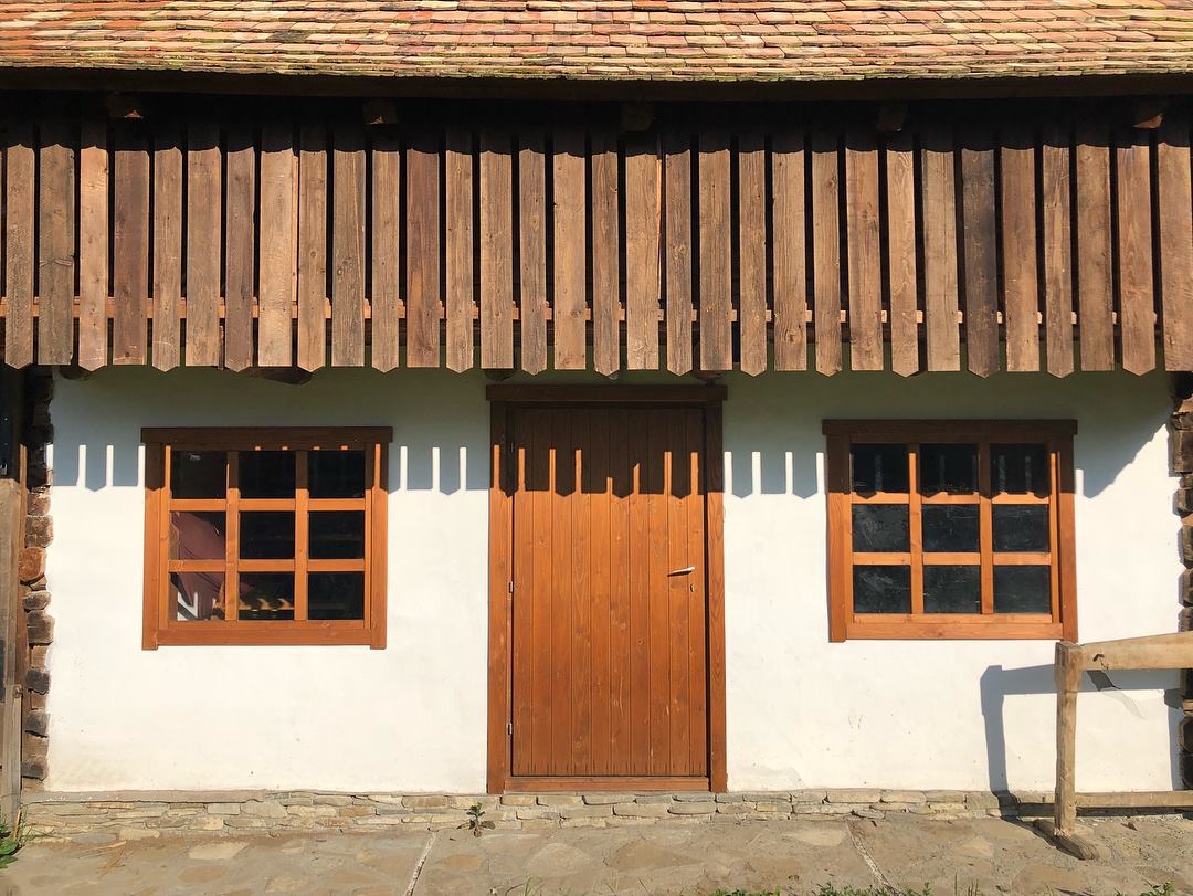 Close-up of the whitewashed building facade with a carved wooden eave, a plank door, and two shuttered windows.