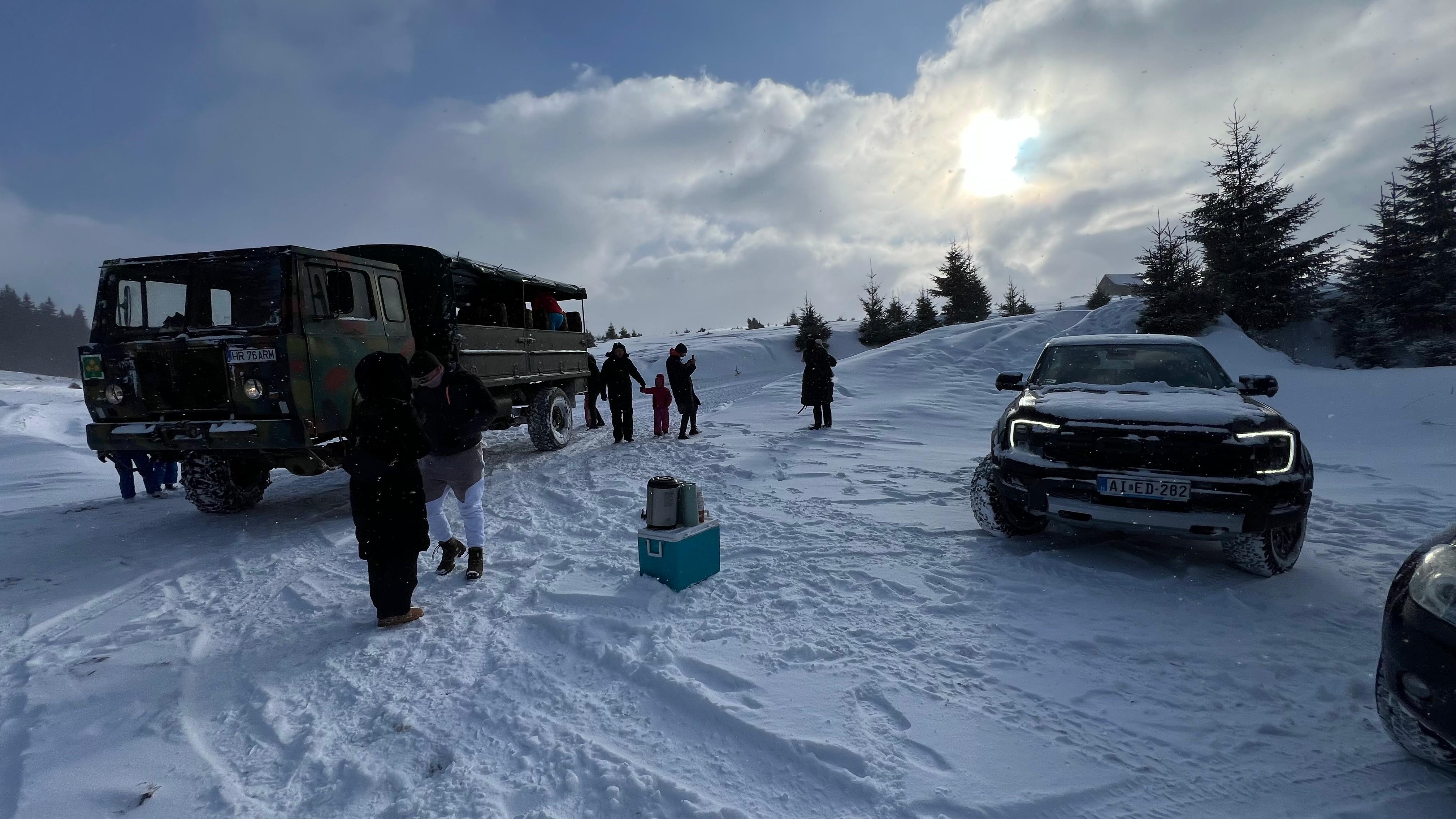 A camouflage-painted off-road tour truck and a pickup truck parked in the snow as visitors gather nearby.