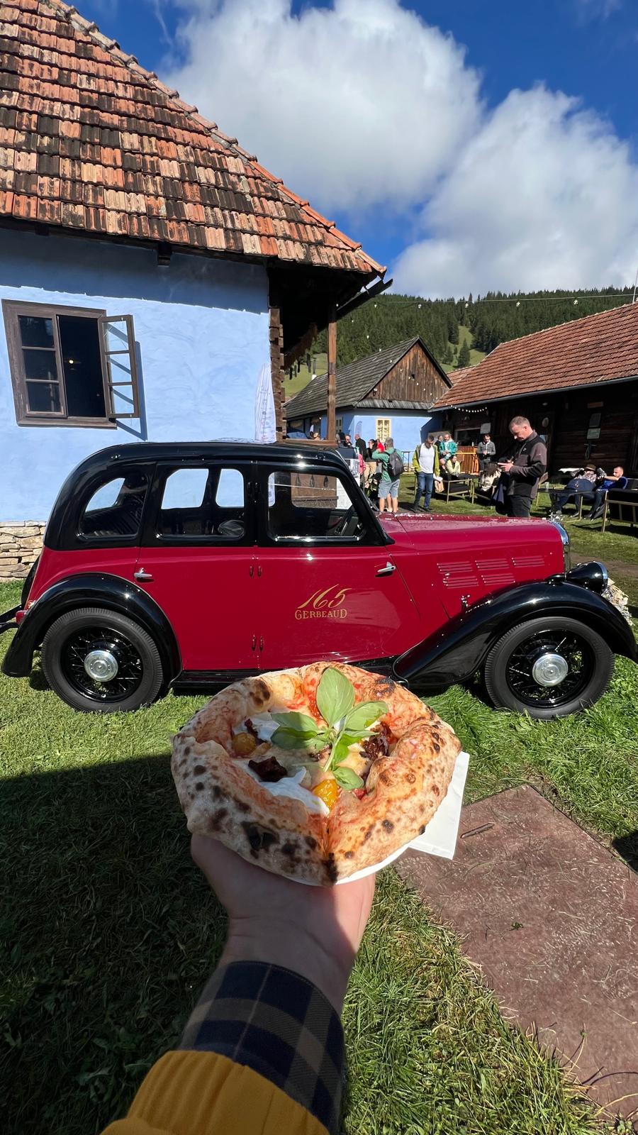Wood-fired pizza held up in front of a vintage red car parked outside a blue-painted house at the festival.