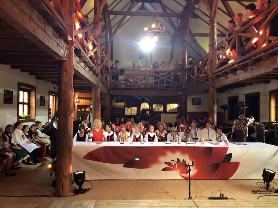 Children in folk costumes perform on stage in the barn, watched by an audience on the floor and mezzanine above.