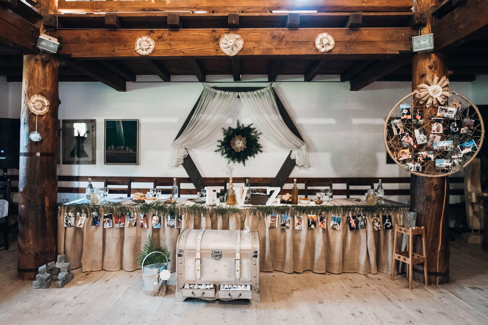 Wedding head table decorated with a lace backdrop, a greenery wreath, and guest photos pinned along its burlap skirt.