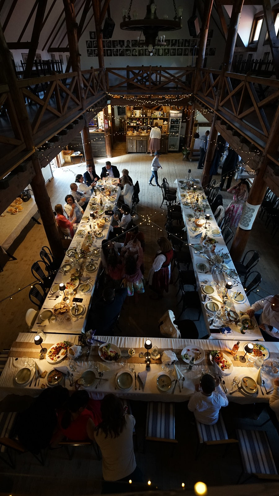 Aerial view of a wedding reception in the barn, guests dining at long tables, children in folk costume nearby, band playing.