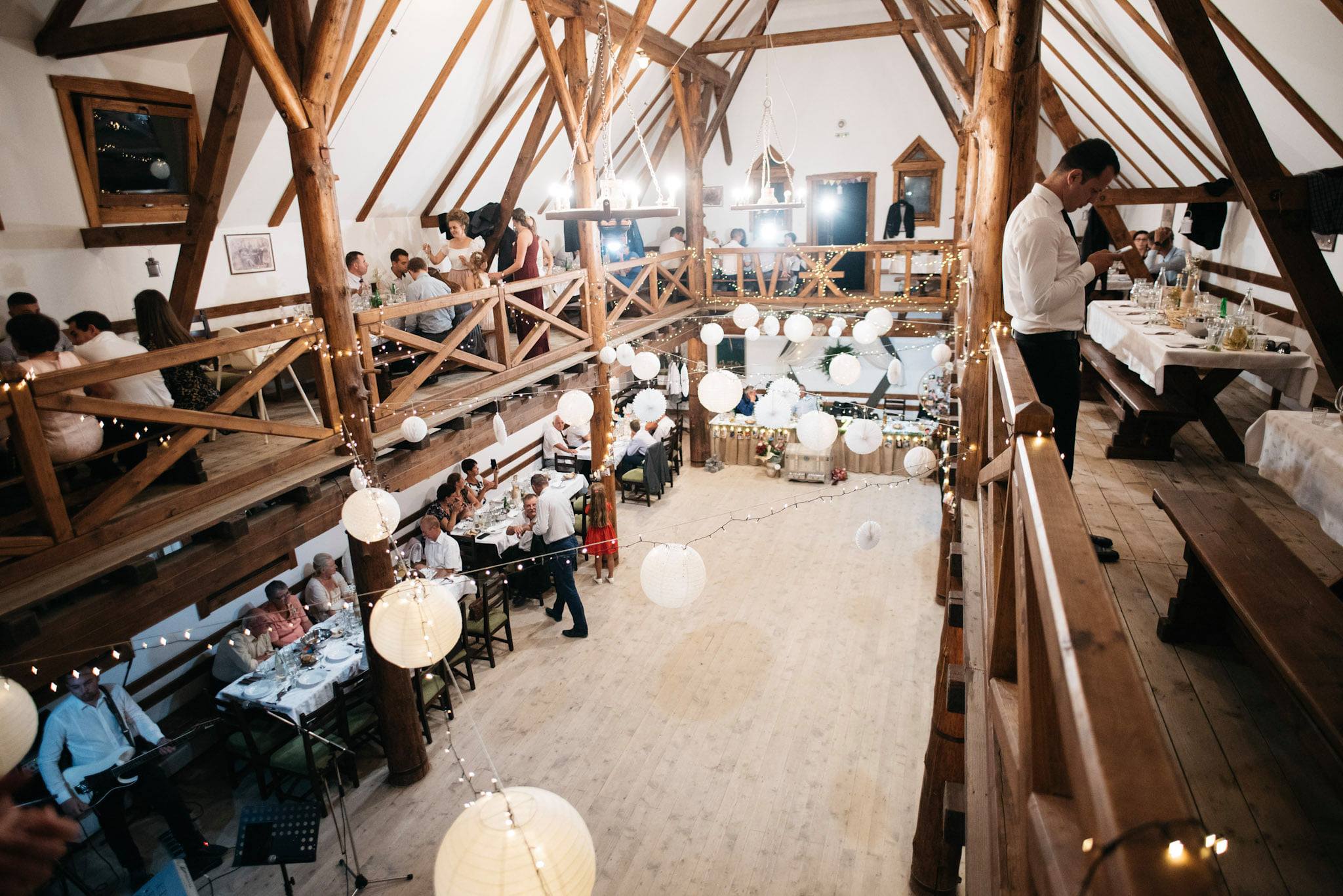 Wedding reception at night in the barn, guests seated at long tables under paper lanterns and string lights, band performing.