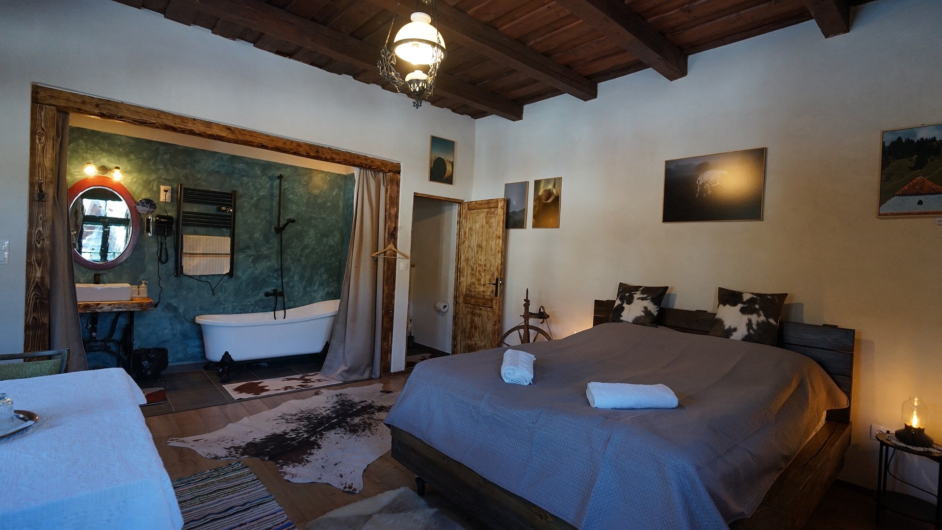 Rustic guesthouse bedroom with a freestanding clawfoot tub, cow-print pillows and rug, and a wood-beamed ceiling.