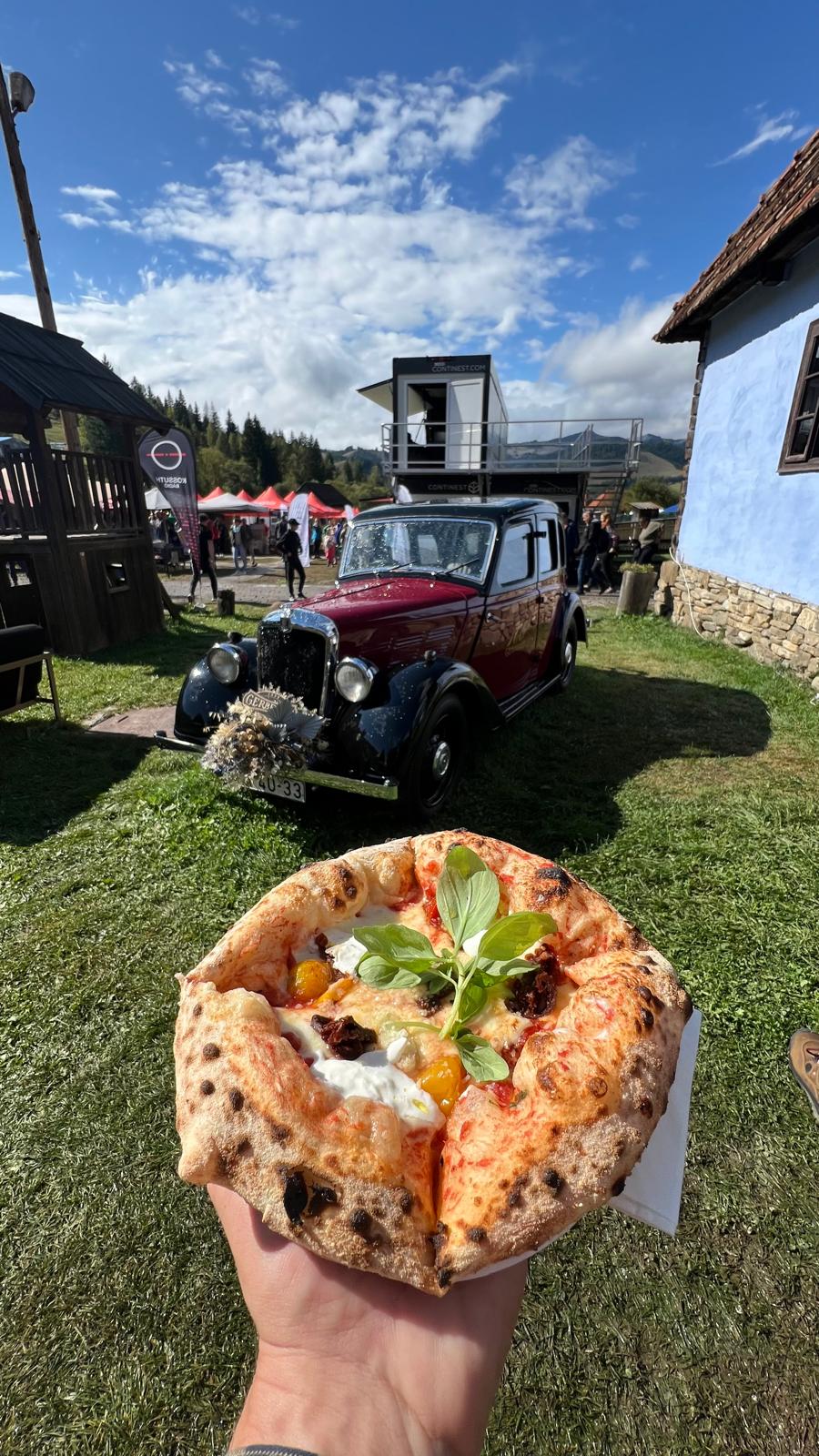 Wood-fired pizza held up in front of a vintage maroon car decorated with flowers, festival tents visible behind.