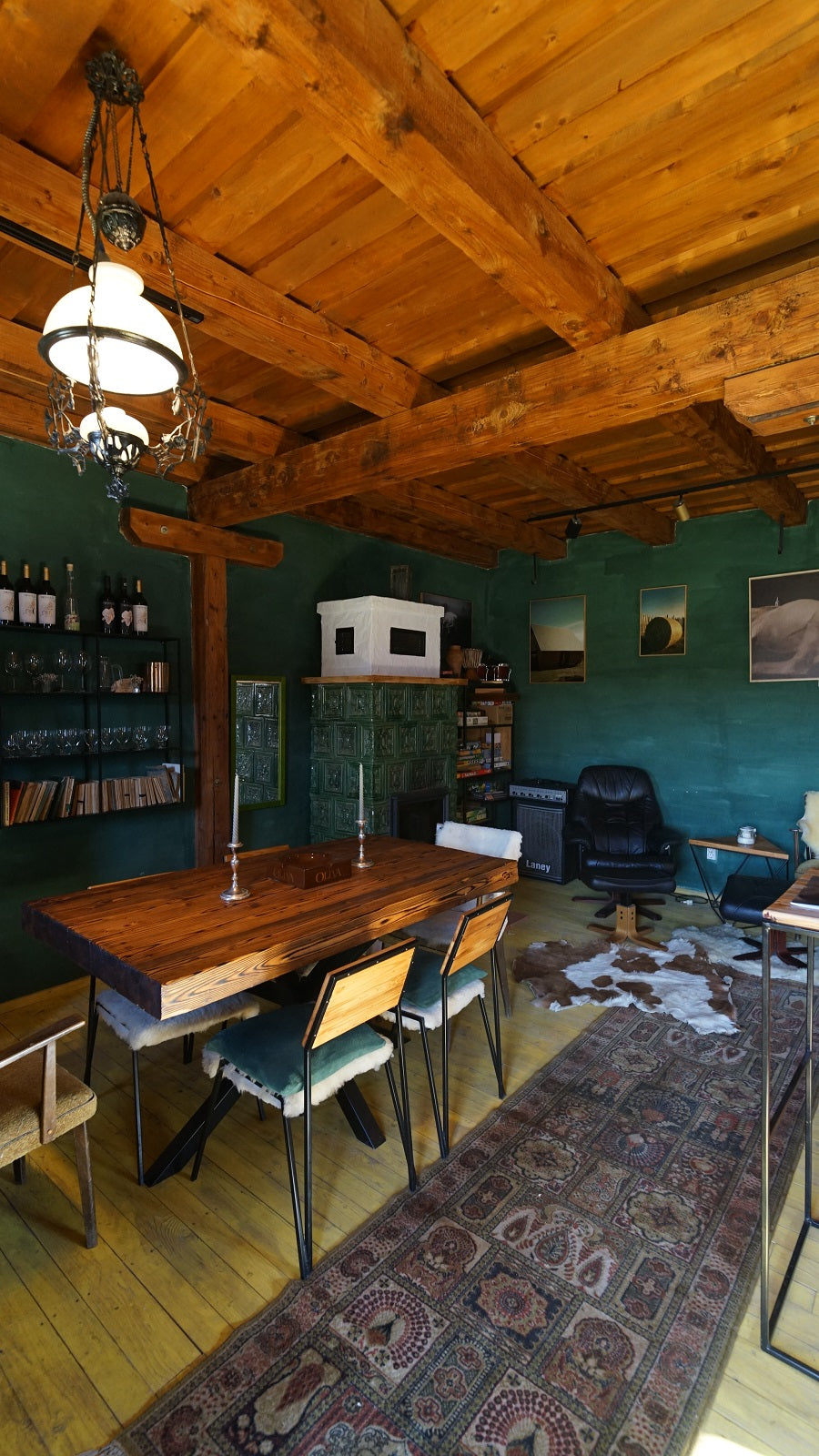 Dining table set with candlesticks, a green tile stove, a wine and glassware shelf, and a cowhide rug on the floor.