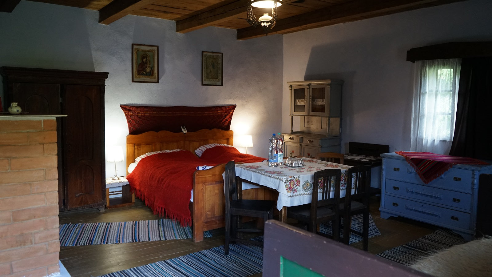 Bedroom with a wood bed draped in a deep red woven blanket, religious icon paintings, and a painted blue dresser.