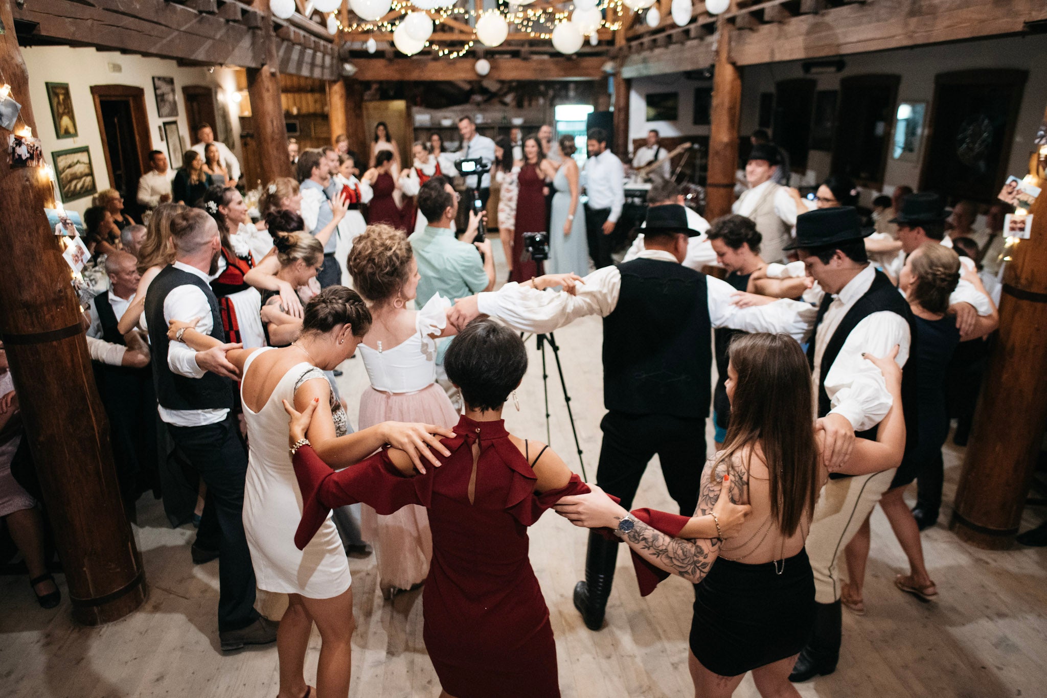 Wedding guests forming a dance circle inside a timber-framed wooden hall illuminated by hanging paper lanterns.